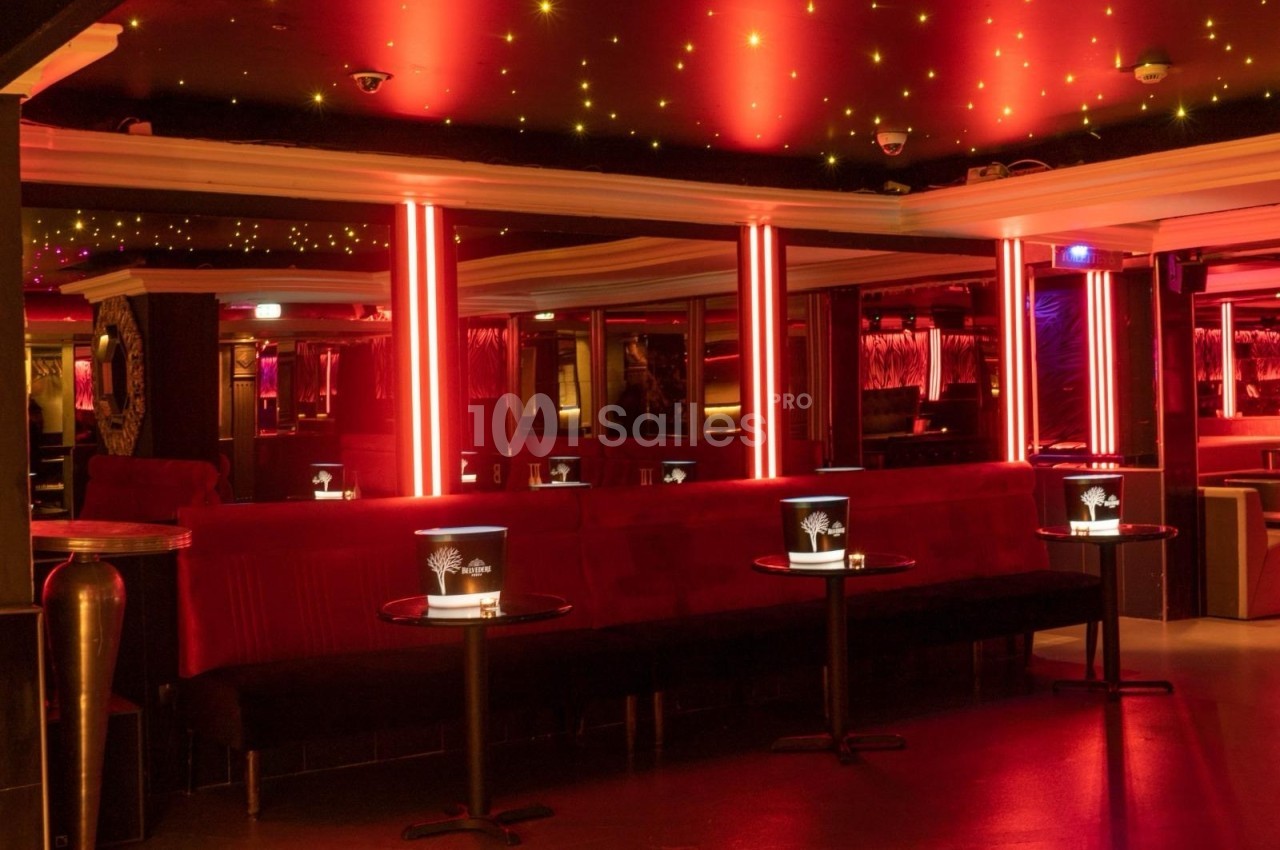 Salle de club avec éclairage rouge, banquettes en velours, tables basses et seaux à champagne.