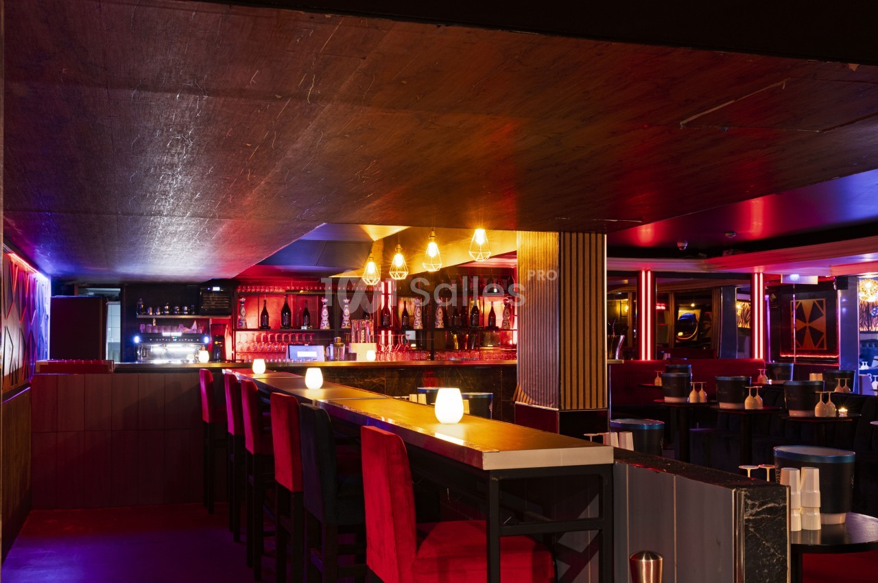 Intérieur d'un bar avec un long comptoir, des tabourets rouges, des lumières tamisées et une ambiance chaleureuse.