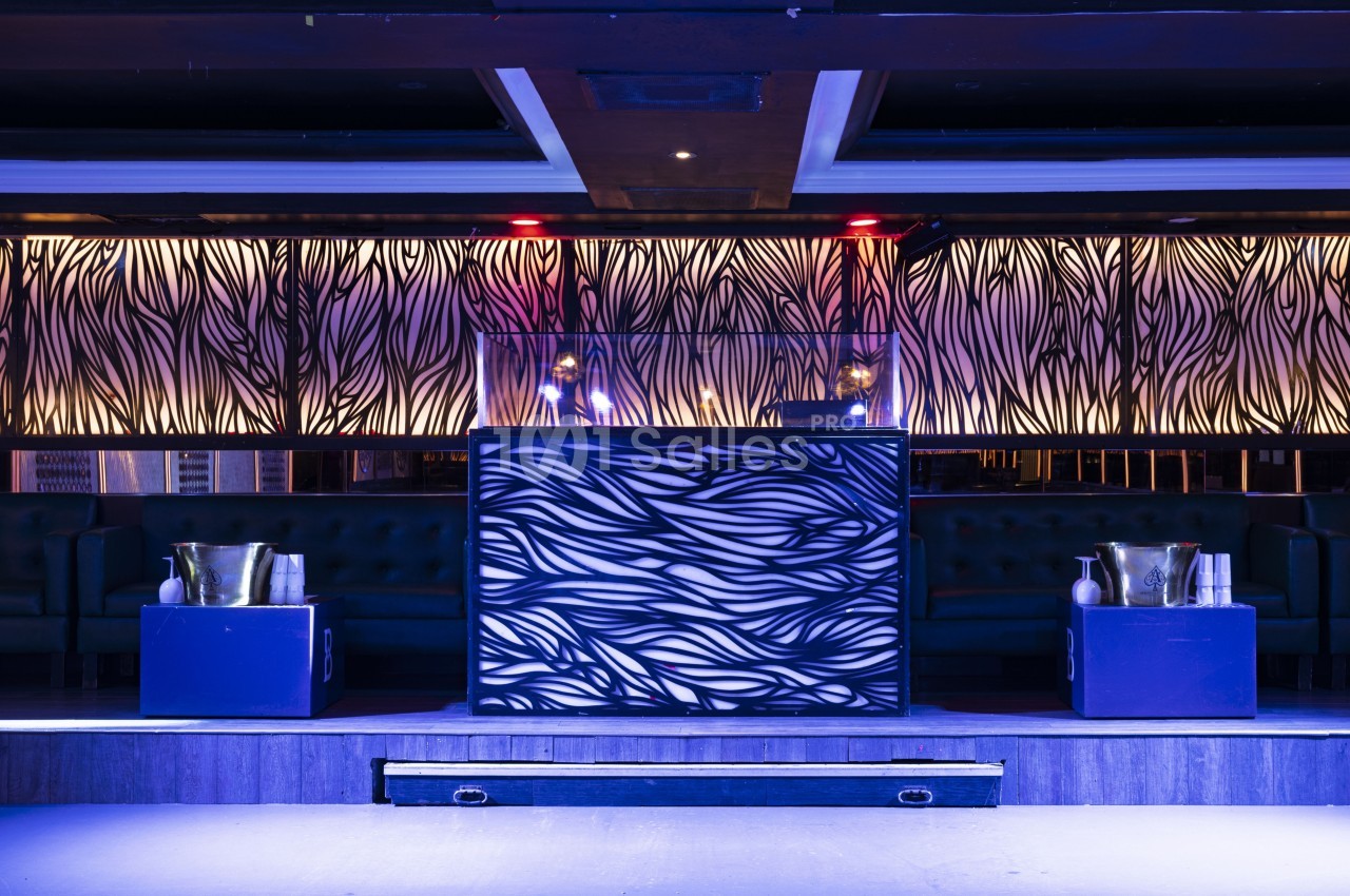 Cabine de DJ avec un design lumineux et abstrait, entourée de banquettes et de seaux à champagne dans une salle sombre.