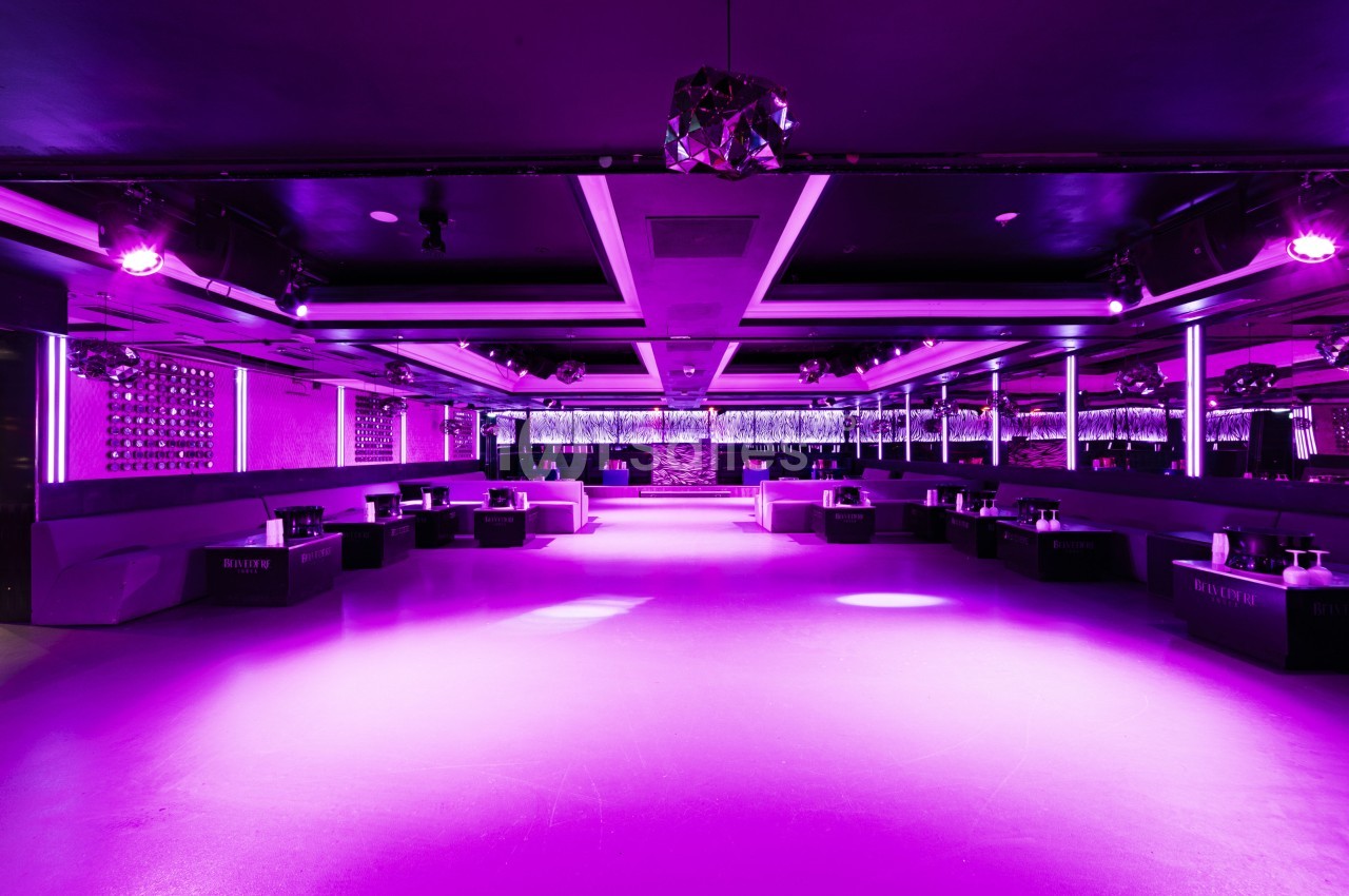 Salle de club vide éclairée par des lumières violettes, avec banquettes et tables disposées le long des murs.
