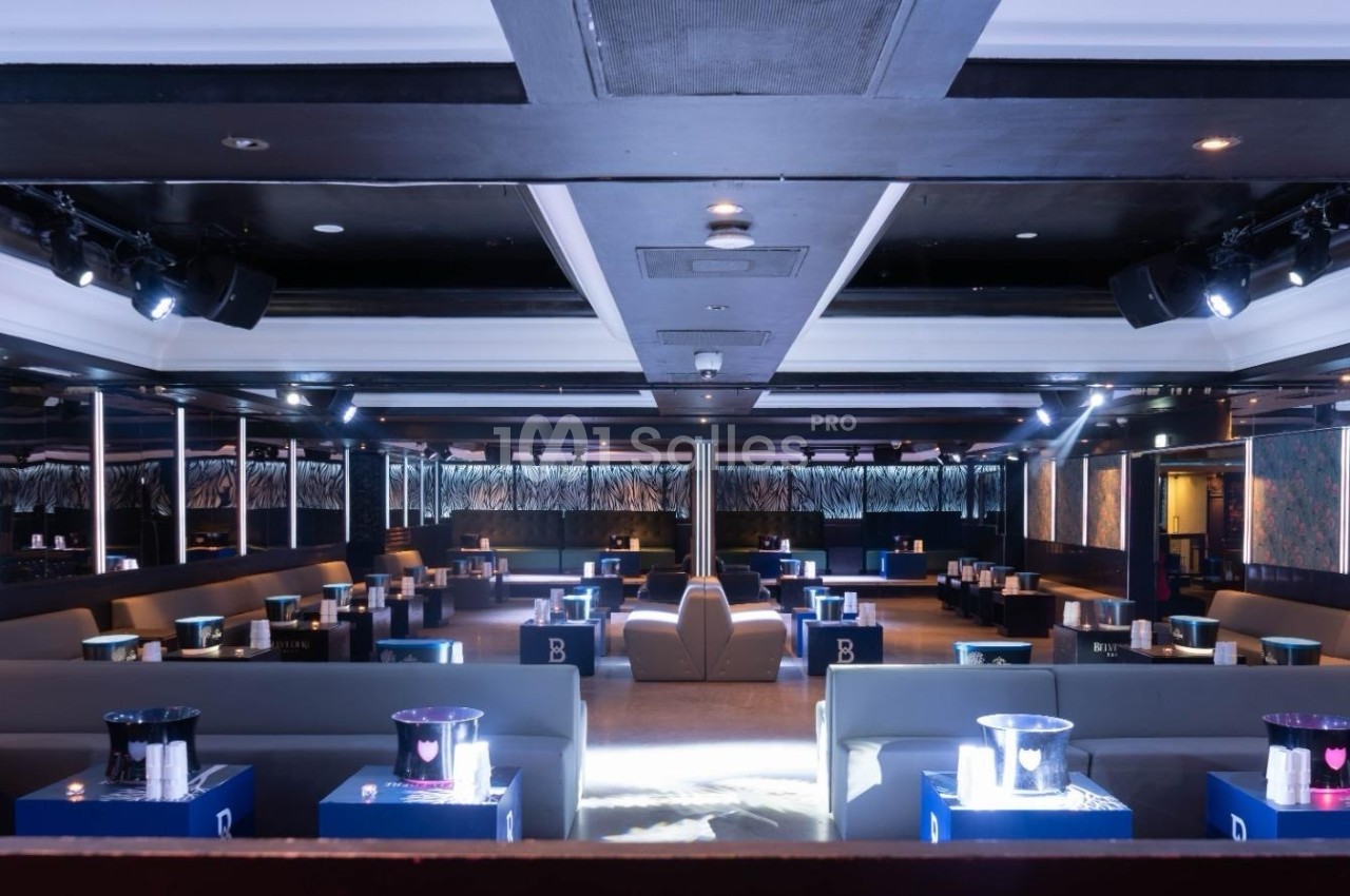 Salle de club avec banquettes grises, tables basses éclairées et ambiance lumineuse tamisée.