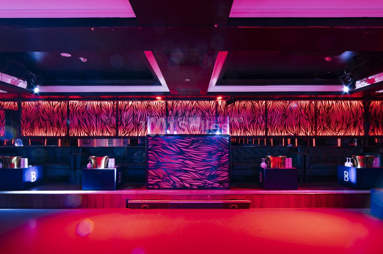 Salle de club avec éclairage rouge et violet, bar central décoré de motifs zébrés et banquettes en arrière-plan.