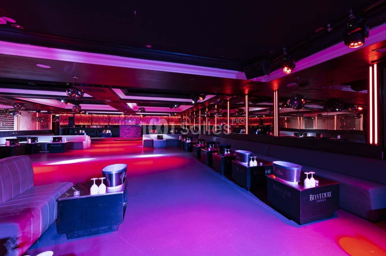 Salle de club vide avec éclairage coloré, banquettes, tables basses et murs ornés de miroirs.