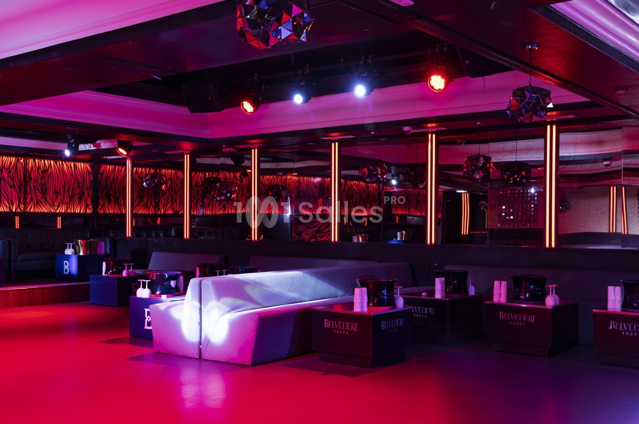 Salle de club avec éclairage rouge et violet, canapés blancs, tables basses et décorations modernes.