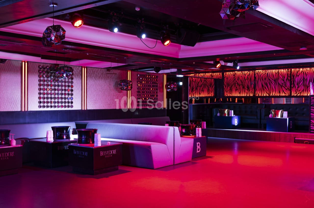 Salle de club avec éclairage rouge, banquettes grises, tables basses et décoration murale moderne.