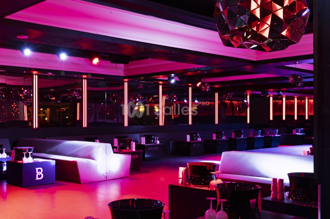 Salle de club avec éclairage rouge et violet, canapés blancs, tables basses et décoration moderne.