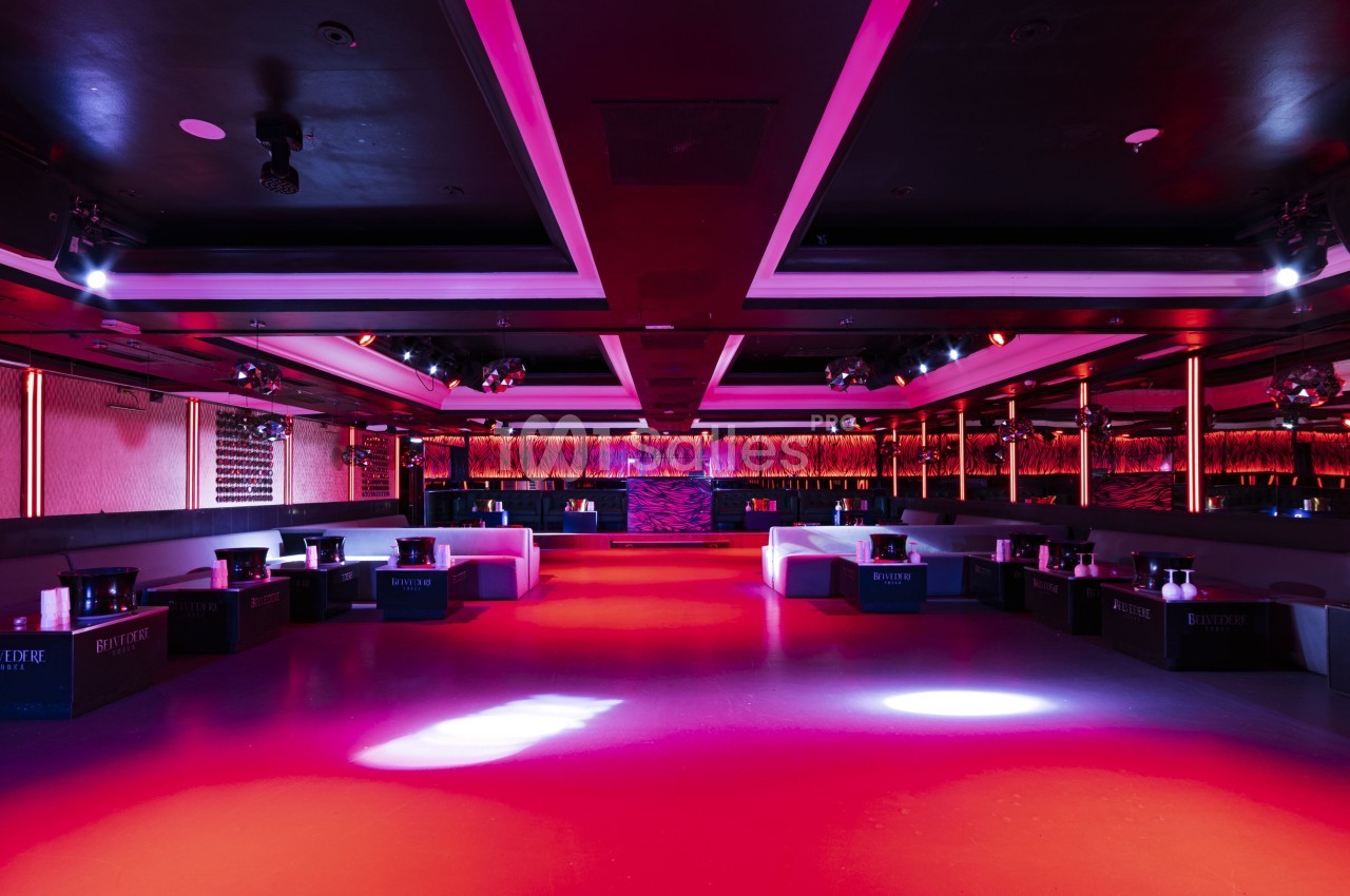 Salle de club vide avec éclairage rose et violet, banquettes alignées sur les côtés et piste centrale dégagée.