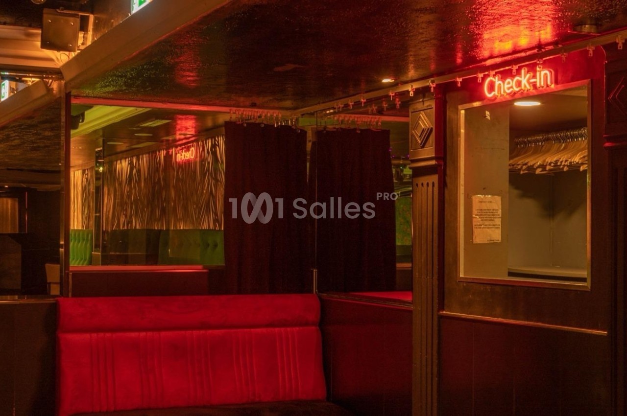 Espace intérieur sombre avec banquette rouge, rideaux noirs et enseigne lumineuse ’Check-in’ au mur.