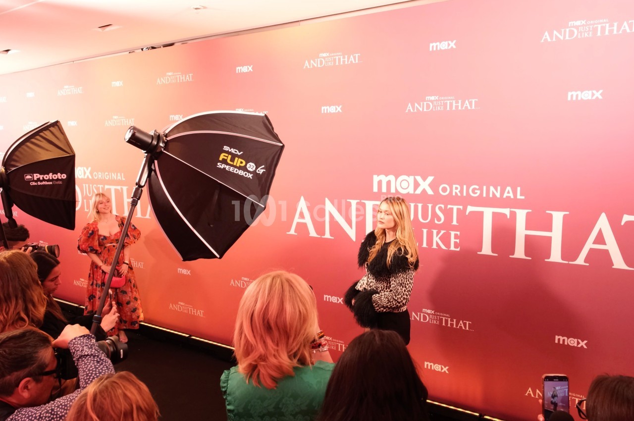Une femme pose sur un tapis rouge devant un mur promotionnel rose, entourée de photographes et de projecteurs.