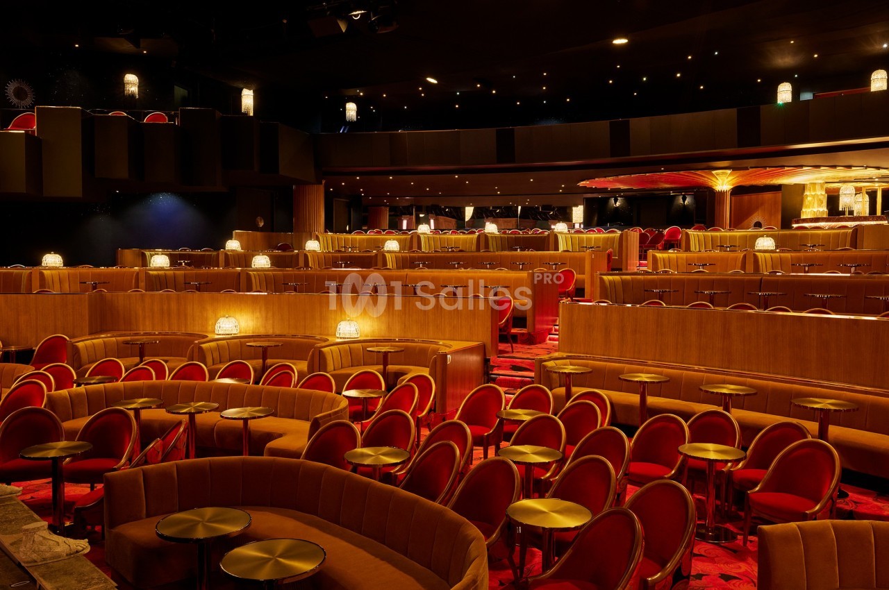 Salle de spectacle avec sièges rouges, tables rondes dorées et éclairage tamisé créant une ambiance chaleureuse.