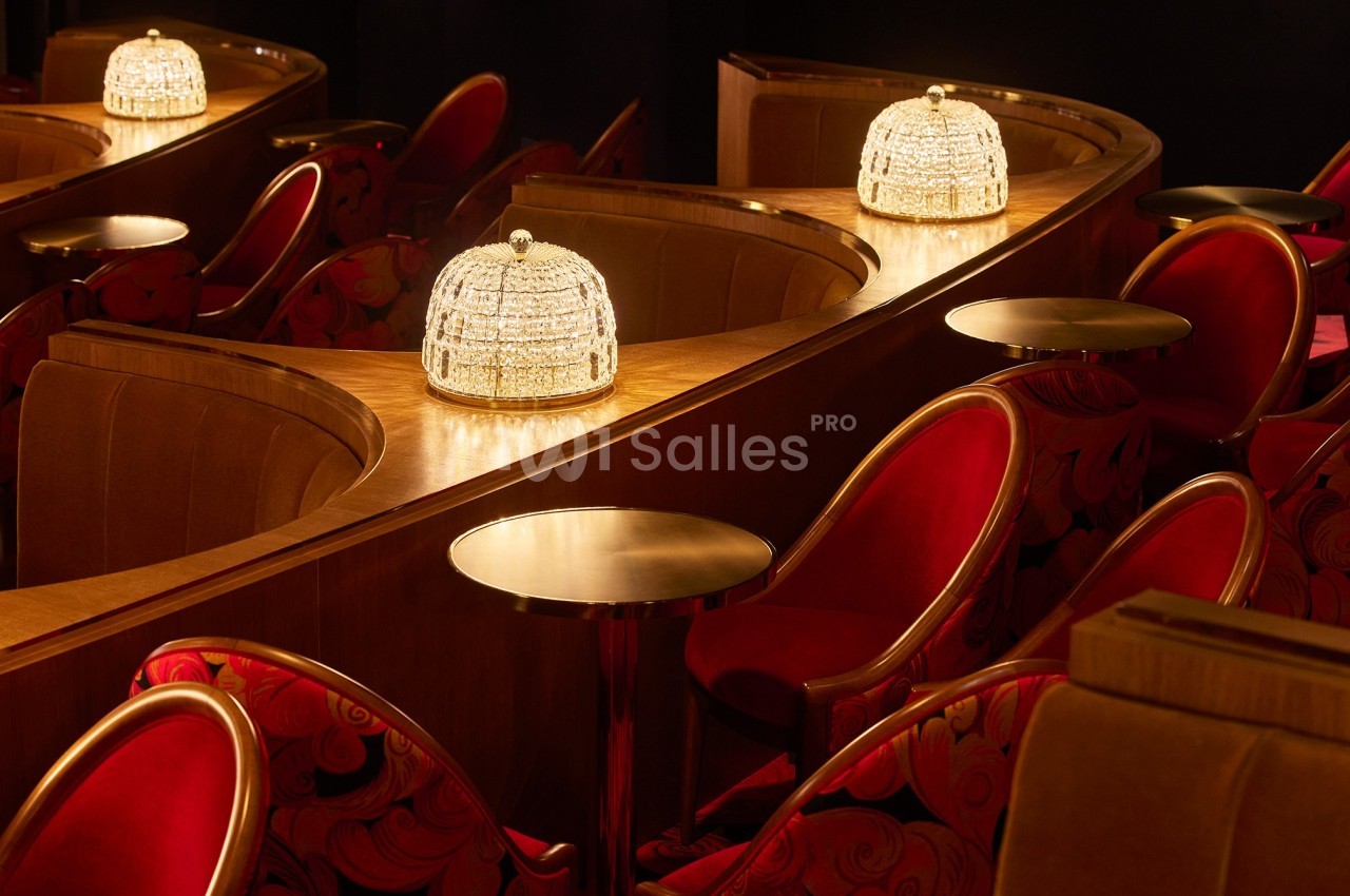 Banquettes en velours rouge, tables rondes en bois et lampes élégantes dans une salle au décor chaleureux.