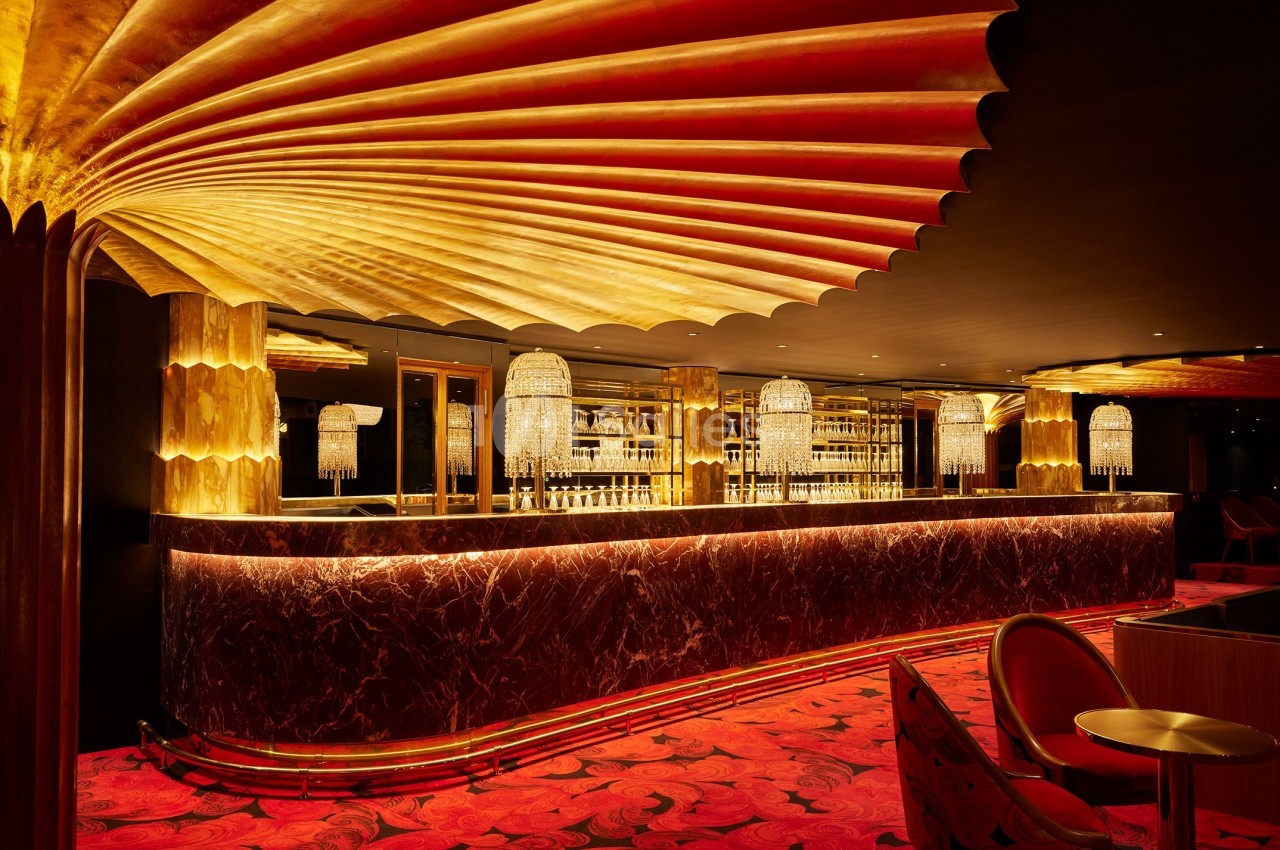 Bar luxueux avec comptoir en marbre, éclairage doré, lustres en cristal et moquette rouge ornée de motifs.