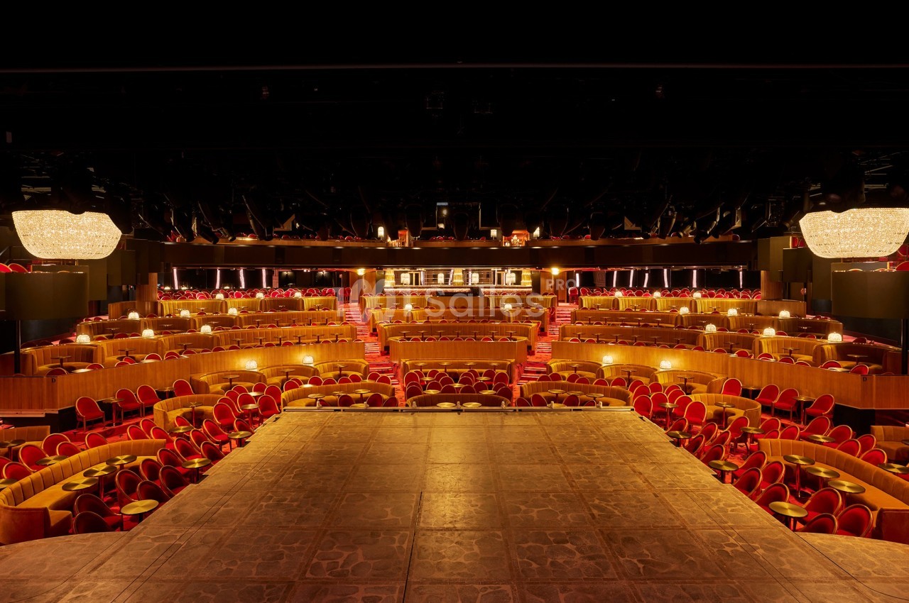 Salle de spectacle avec scène au premier plan, tables et fauteuils rouges disposés en gradins sous des lustres.