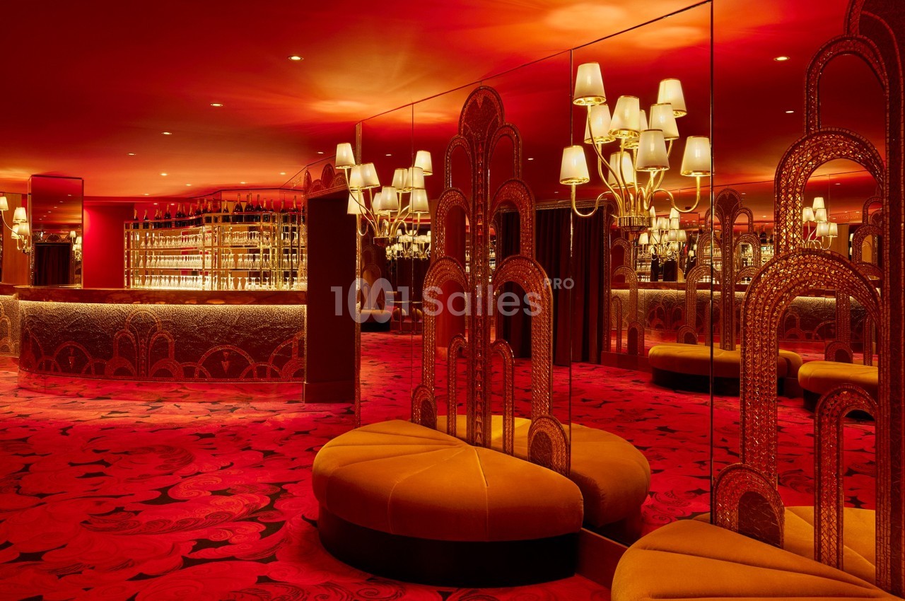 Salon luxueux avec éclairage rouge, banquettes arrondies, miroirs, lustres et un bar en arrière-plan.