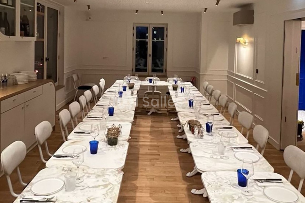 Salle à manger lumineuse avec deux longues tables blanches, chaises alignées, verres bleus et vaisselle disposée.
