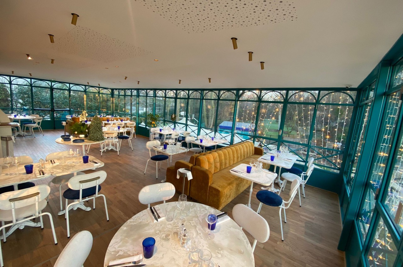 Salle de restaurant lumineuse avec grandes baies vitrées, tables dressées, chaises blanches et vue sur un espace extérieur.