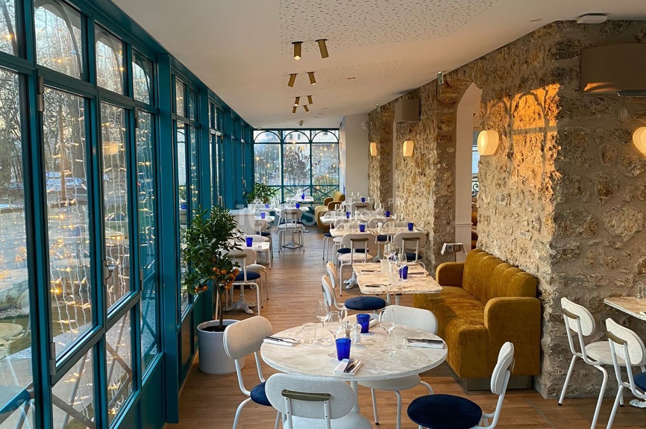 Salle de restaurant lumineuse avec murs en pierre, grandes baies vitrées, tables dressées et chaises blanches.