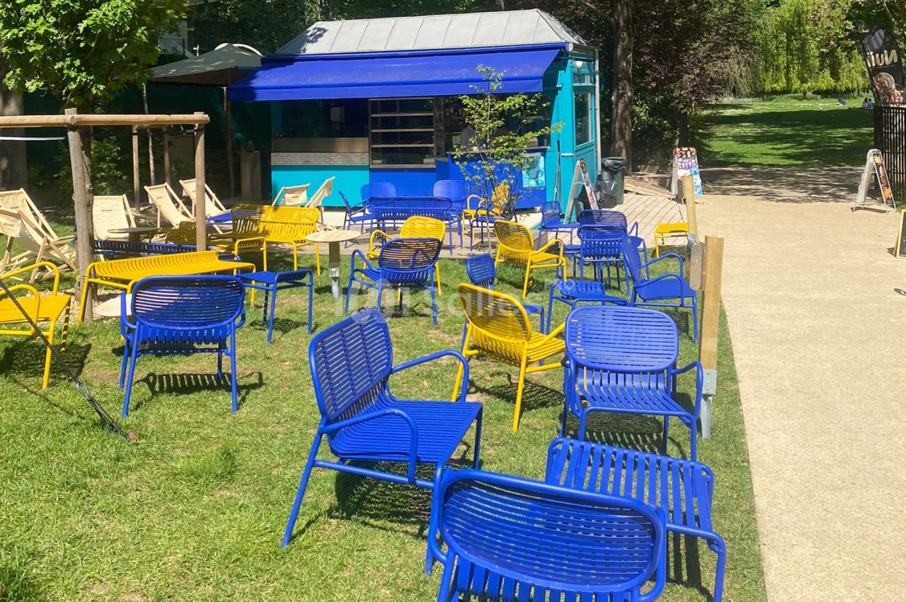 Chaises bleues et jaunes disposées sur une pelouse devant un kiosque bleu, entouré d'arbres et d'un chemin pavé.