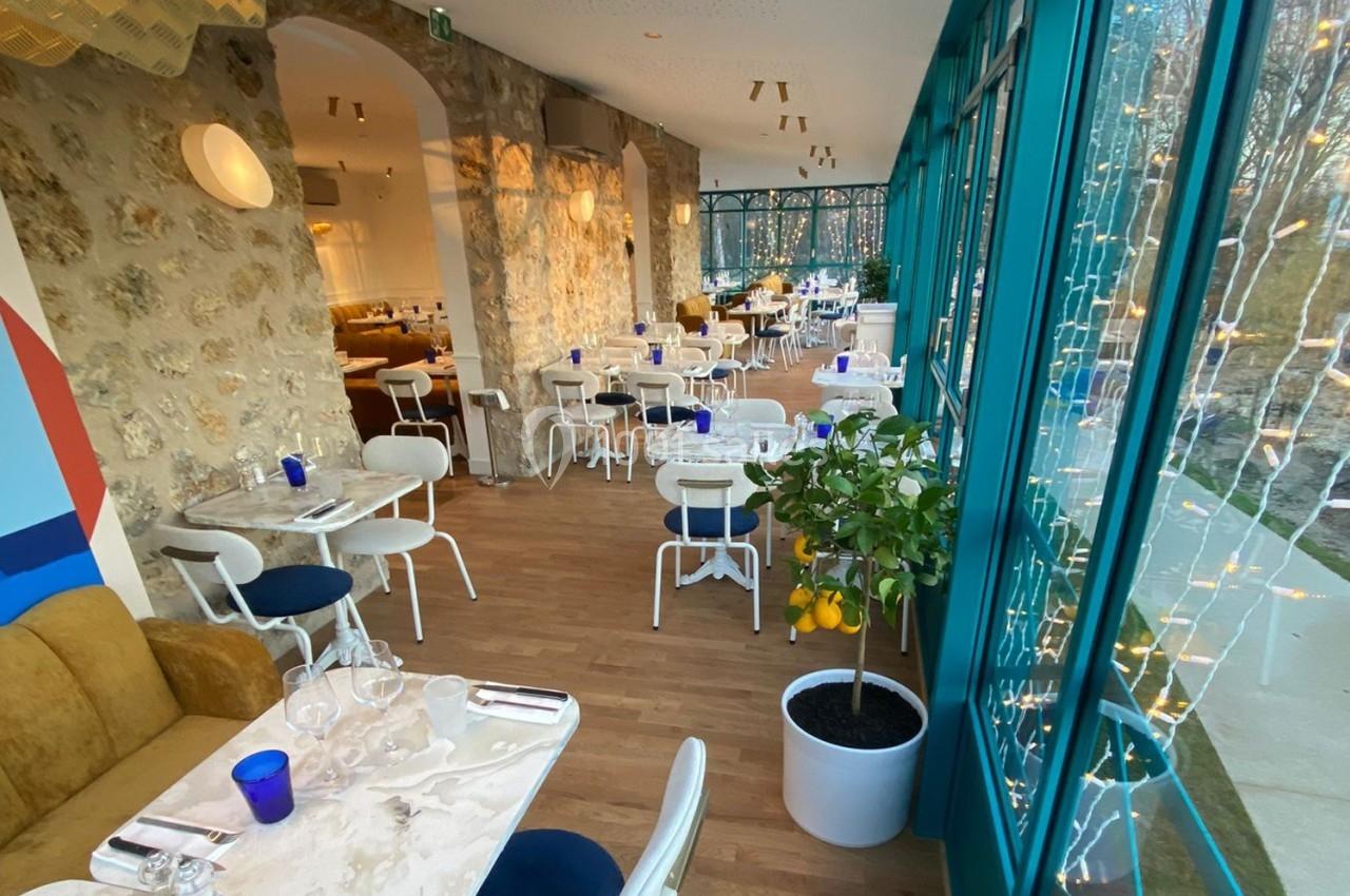 Salle de restaurant lumineuse avec murs en pierre, grandes baies vitrées, tables dressées et un citronnier en pot.