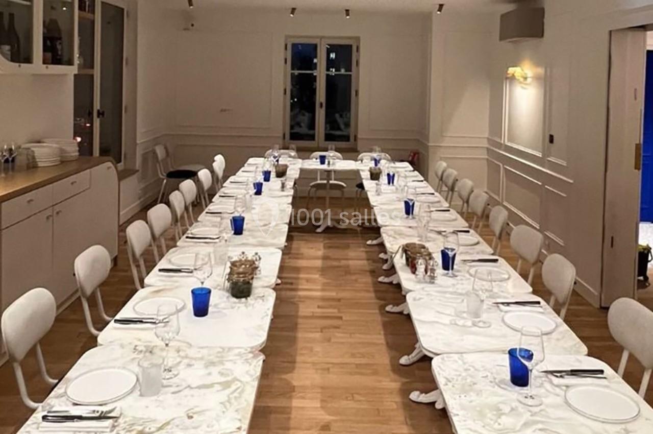 Salle à manger lumineuse avec deux longues tables en marbre blanc, dressées avec des verres bleus et des couverts.