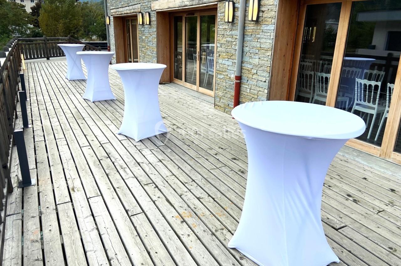 Tables hautes recouvertes de nappes blanches sur une terrasse en bois, avec des chaises visibles à travers les baies vitrées.