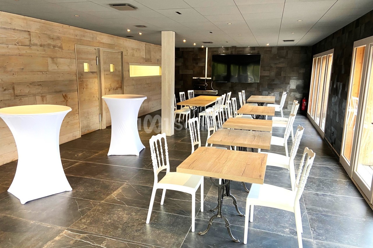 Salle lumineuse avec tables en bois, chaises blanches, mange-debout et grandes fenêtres laissant entrer la lumière naturelle.