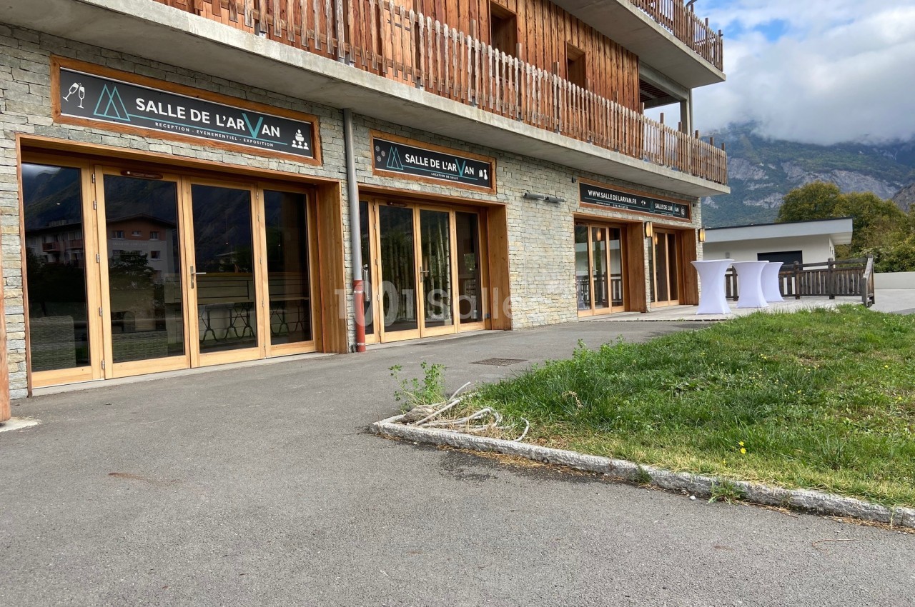 Façade d'un bâtiment en bois et pierre avec des baies vitrées et des enseignes indiquant ’Salle de l'Arvan’.
