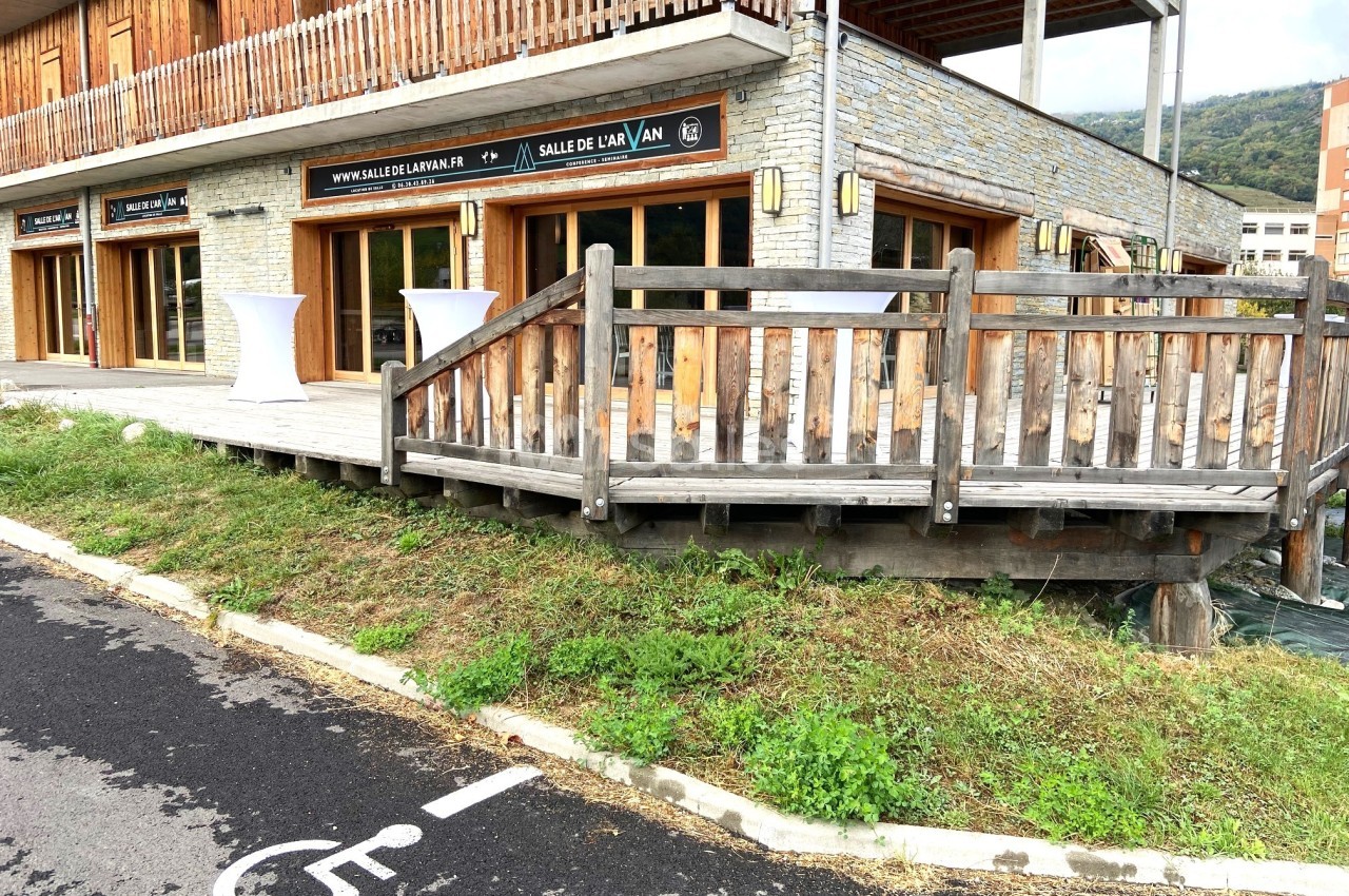 Rampe en bois menant à l'entrée d'un bâtiment avec façade en pierre et bois, vue depuis un parking avec place PMR.