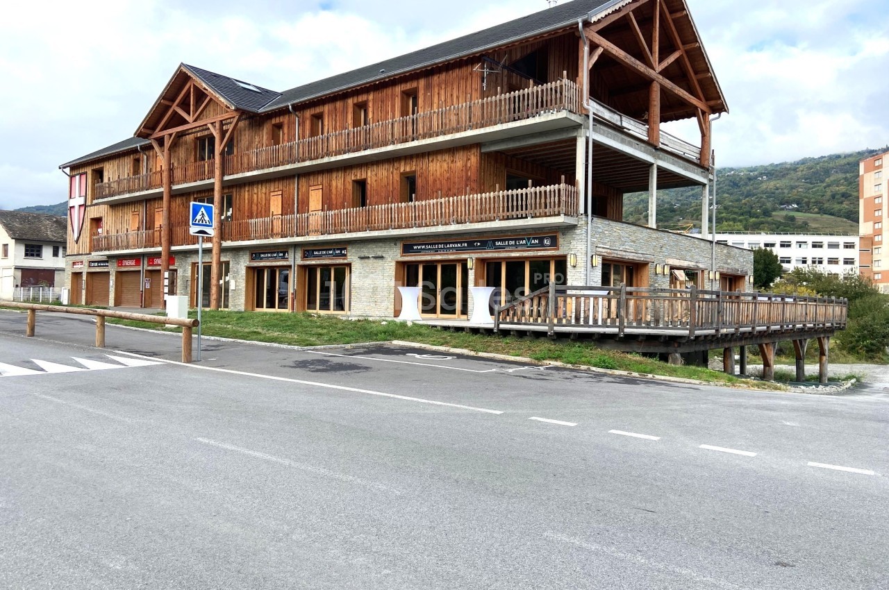 Bâtiment en bois et pierre à deux étages avec balcons, situé près d'une route et entouré de montagnes.