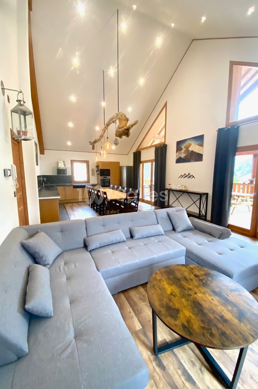 Salon spacieux avec canapé d'angle gris, table basse ronde en bois, cuisine ouverte et grande baie vitrée.