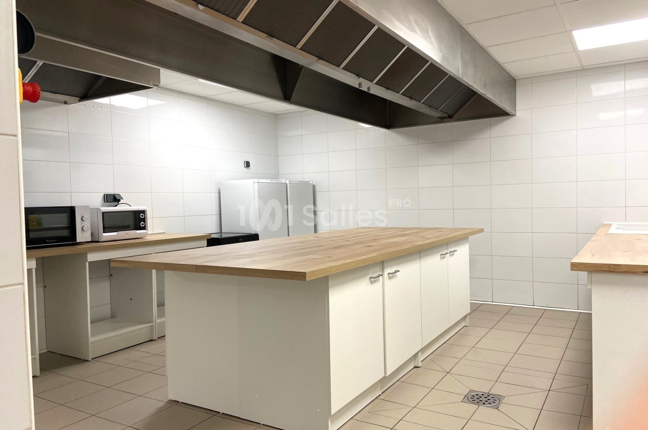 Cuisine professionnelle avec plan de travail central, électroménagers et hotte en acier inoxydable dans un espace carrelé.