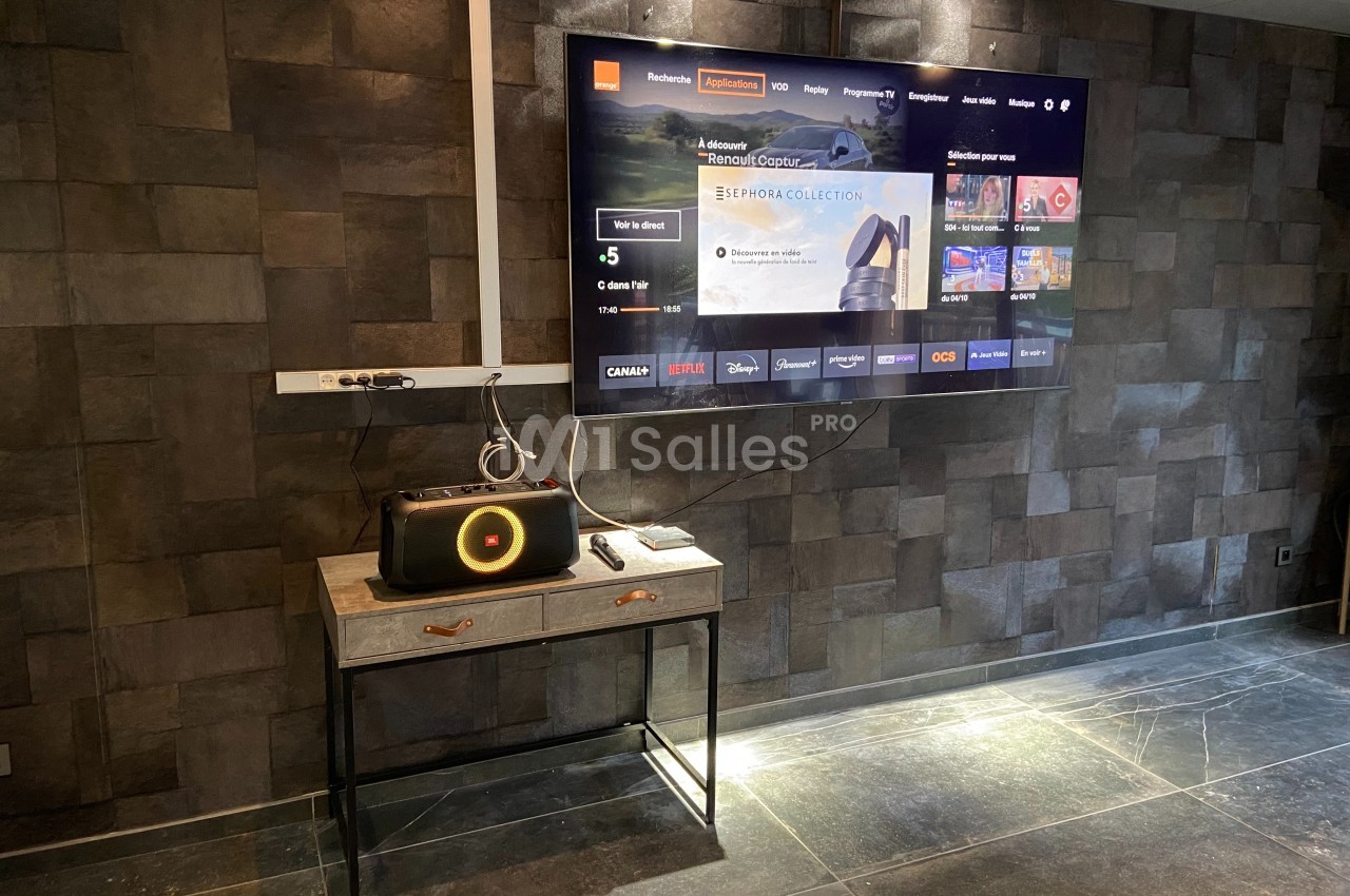 Télévision murale affichant une interface de streaming, avec une enceinte posée sur une table dans une pièce sombre.