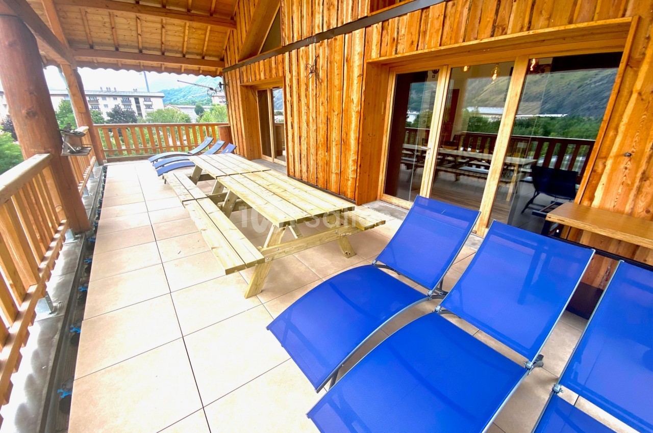 Terrasse en bois avec table de pique-nique et chaises longues bleues, offrant une vue sur des montagnes et des bâtiments.