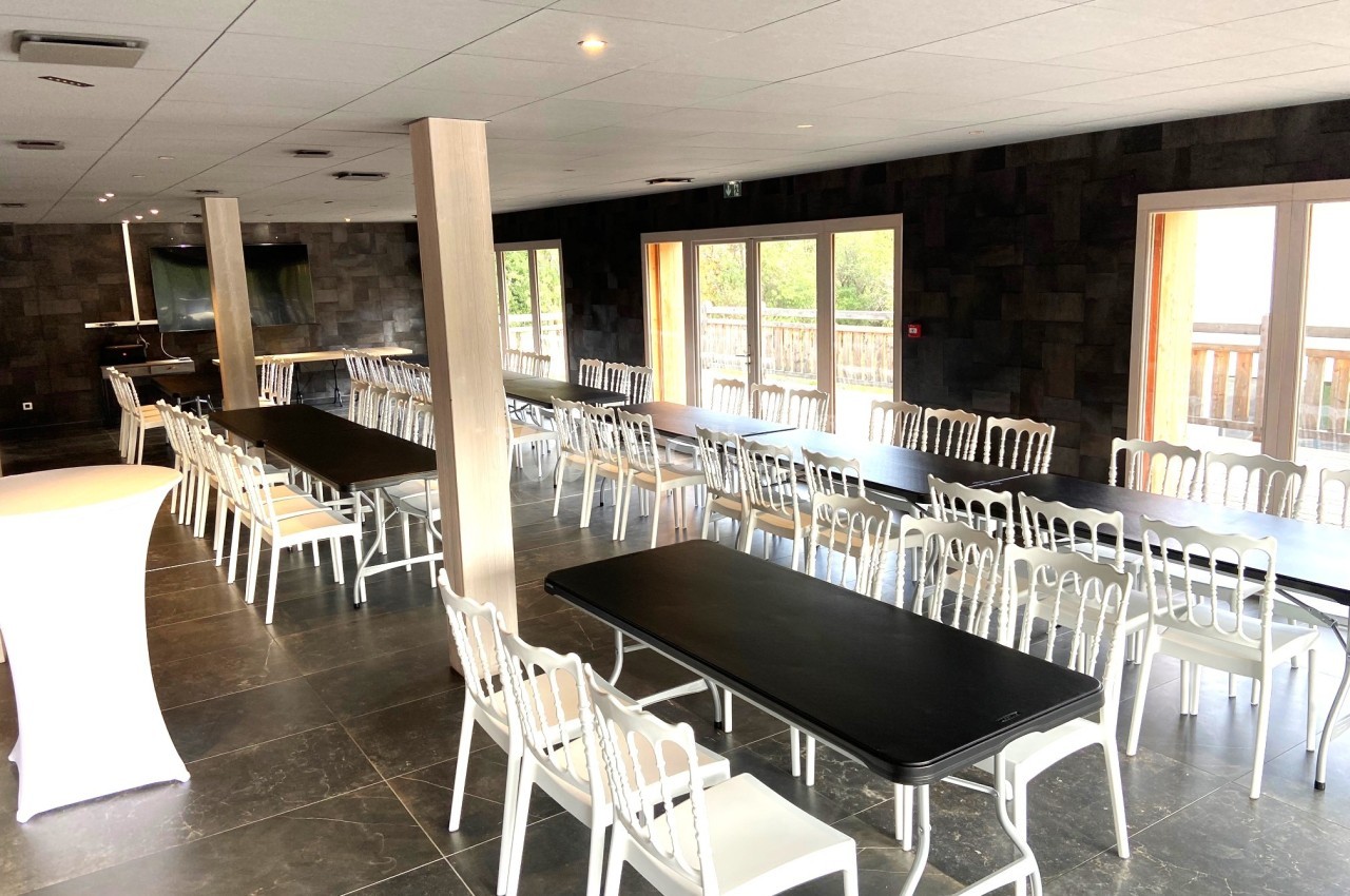 Salle lumineuse avec tables noires alignées, chaises blanches et grandes baies vitrées donnant sur un balcon.
