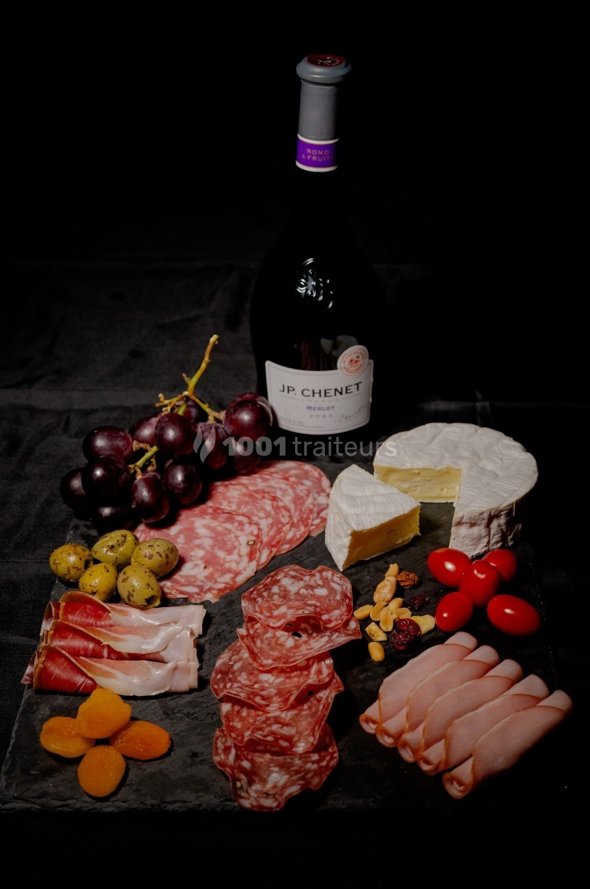 Plateau de charcuterie avec fromages, fruits, olives et une bouteille de vin rouge sur fond noir.