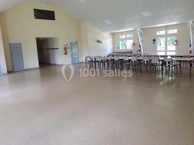 Salle polyvalente vide avec des chaises et des tables alignées près des fenêtres, décorée de ballons tricolores.
