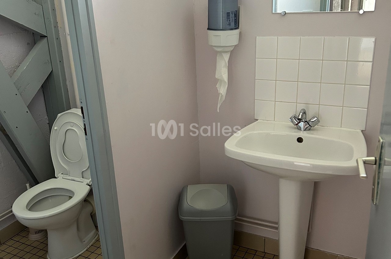 Toilettes avec lavabo, miroir, distributeur de papier, poubelle et carrelage au sol dans un espace restreint.