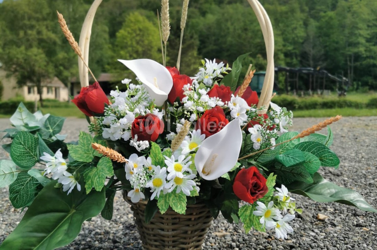 Panier en osier rempli de roses rouges, arums blancs, marguerites et feuillage, posé sur un sol gravillonné en extérieur.