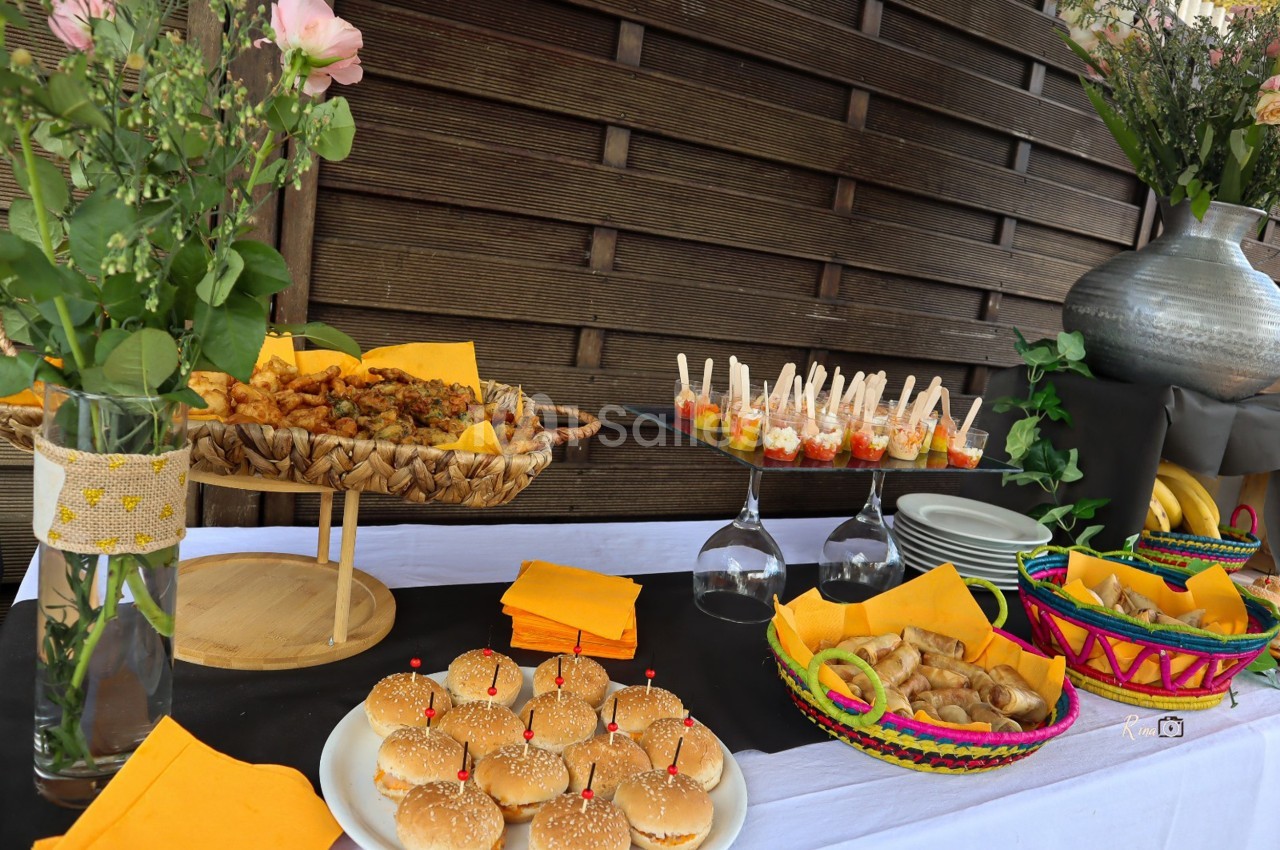 Buffet coloré avec mini-burgers, amuse-bouches, fleurs et paniers garnis, disposé sur une table en extérieur.