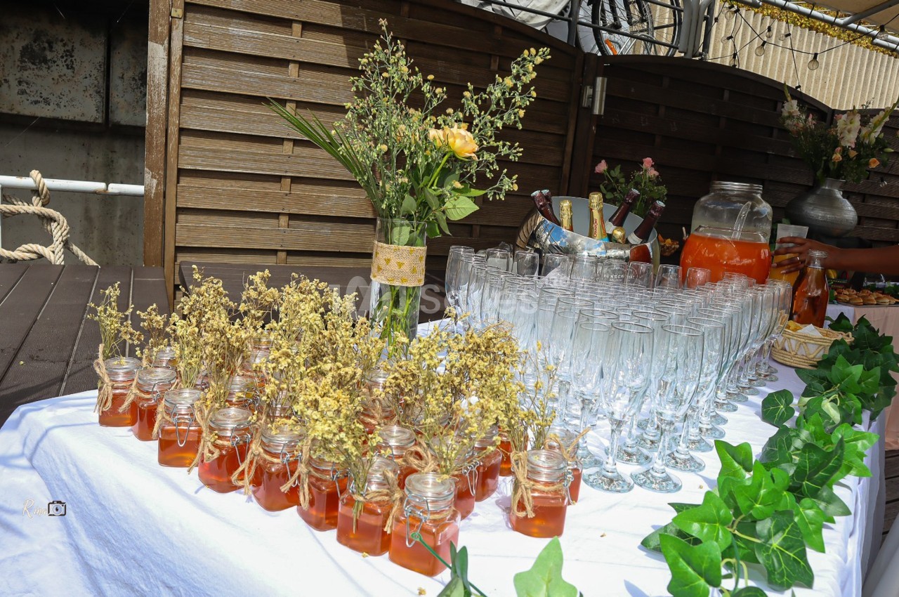Table décorée avec des pots de miel, des fleurs séchées, des verres à pied et des boissons pour un événement en extérieur.