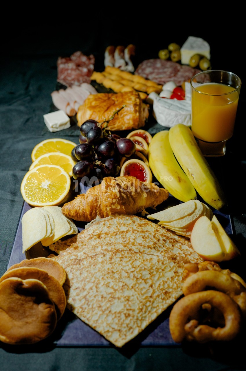 Assortiment de viennoiseries, fruits, charcuteries, fromage et jus d'orange sur une table sombre.