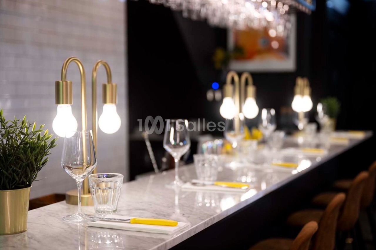 Comptoir en marbre avec couverts, verres et lampes design, entouré de tabourets dans un cadre élégant.