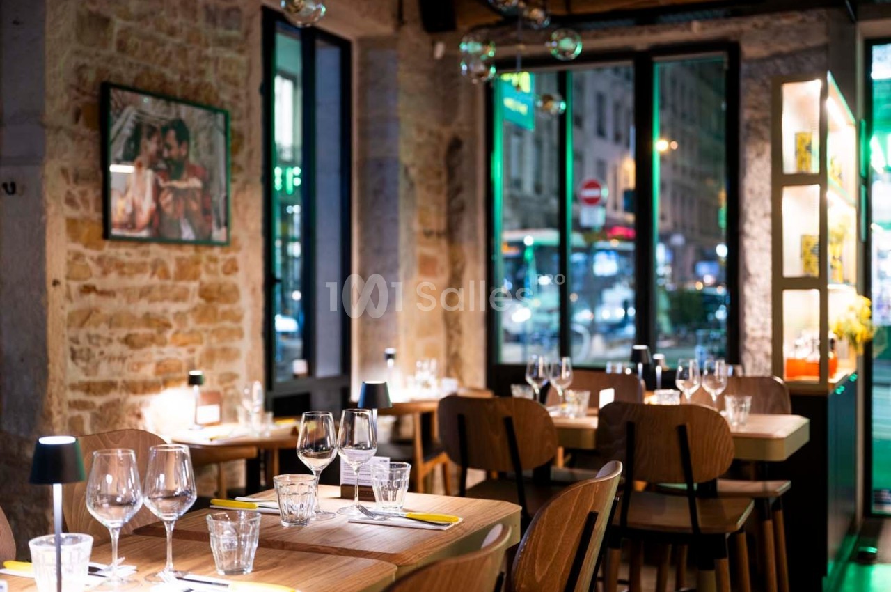 Salle de restaurant chaleureuse avec murs en pierre, tables dressées et vue sur une rue animée à travers de grandes fenêtres.