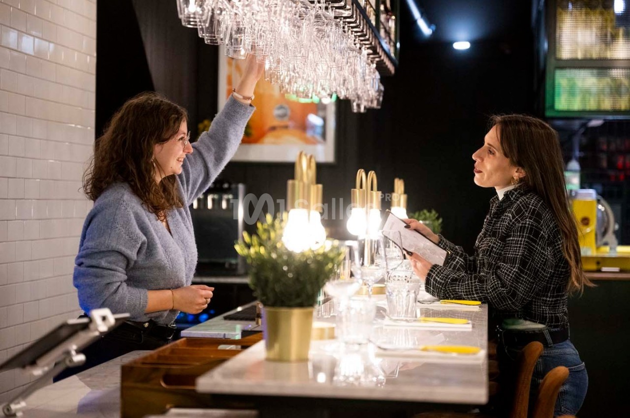 Deux femmes discutent au comptoir d'un bar, l'une tendant la main vers des verres suspendus.