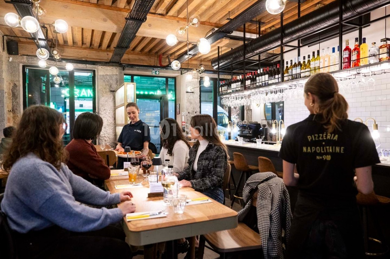 Des clients discutent autour de tables en bois dans une pizzeria animée avec un décor moderne et chaleureux.