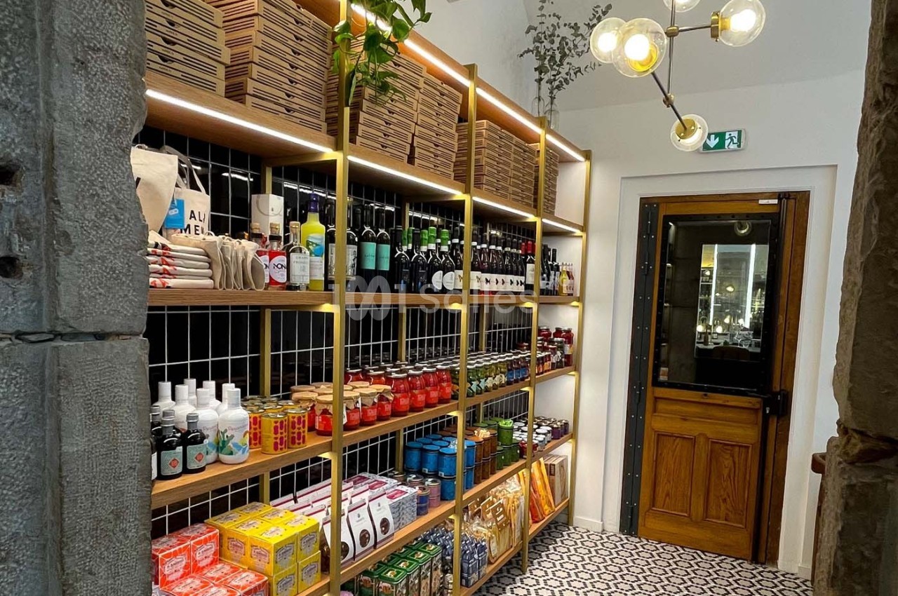 Étagères en bois remplies de produits alimentaires et ménagers dans une petite épicerie lumineuse avec sol carrelé.