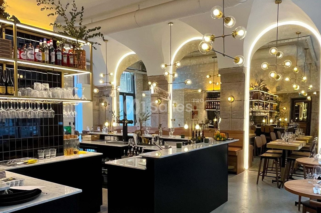 Intérieur d'un restaurant moderne avec arches lumineuses, bar central, étagères de bouteilles et tables dressées.