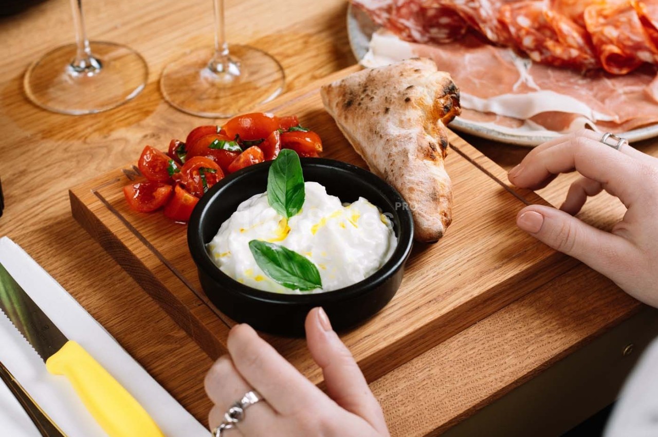 Assiette en bois avec burrata garnie de basilic, tomates cerises, pain et charcuterie en arrière-plan.