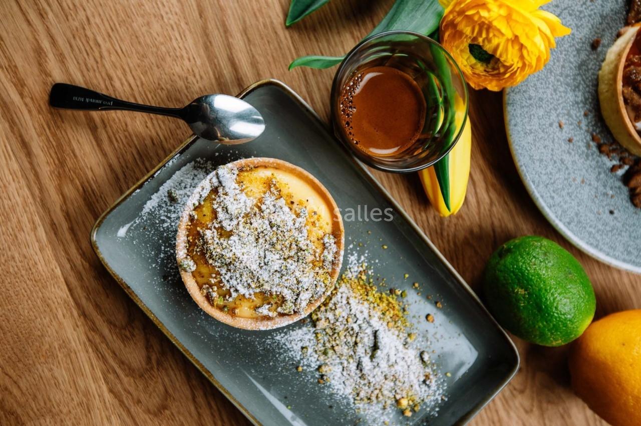 Tartelette saupoudrée de sucre glace sur une assiette, accompagnée d'un espresso et de fruits sur une table en bois.