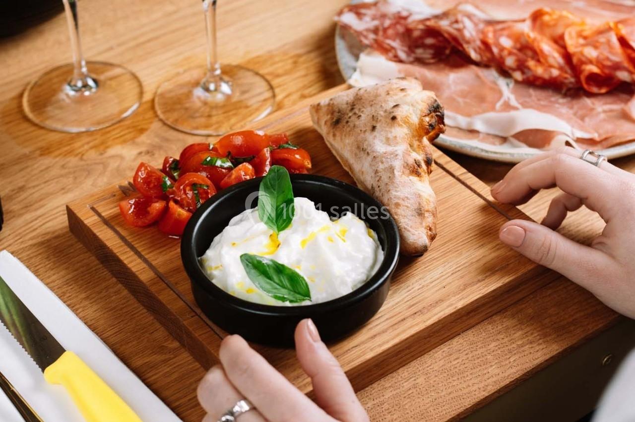 Planche en bois avec burrata, tomates cerises, basilic, pain et charcuterie en arrière-plan.
